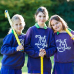 3 girls holding hockey sticks looking at the camera and smiling, sanding on a hockey field.