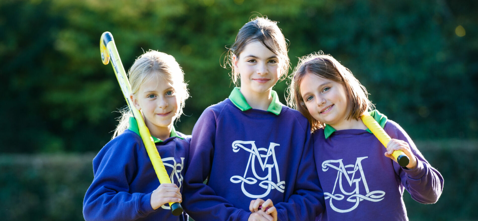 3 girls holding hockey sticks looking at the camera and smiling, sanding on a hockey field.